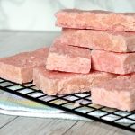 A stack of pink coconut ice bars, reminiscent of a cherished coconut ice recipe, rests on a black wire rack. The unevenly layered bars boast a rough texture, while beneath them lies a colourful striped cloth on a light wooden surface.
