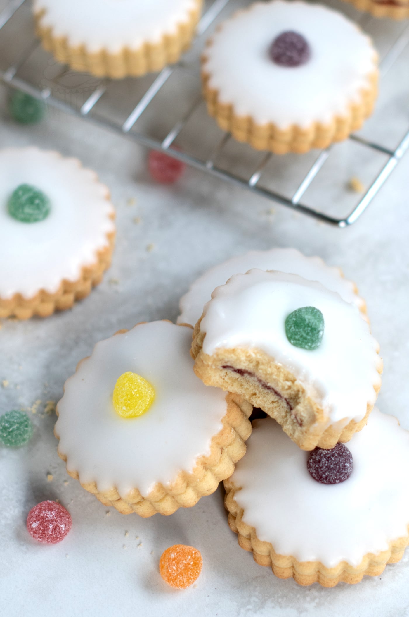 Empire biscuits, iced cookies with colourful jelly tot toppings sit on a cooling rack and countertop. One cookie shows a bite taken out, revealing a jam filling. Scattered jelly sweets surround these tempting treats.