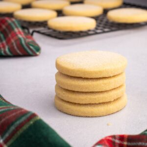 Stack of round Scottish shortbread biscuits surrounded by tartan fabric, with more cooling on a wire rack behind.