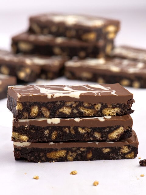 Stack of Scottish chocolate tiffin slices in focus, with a swirled chocolate topping and scattered biscuit pieces in the background.