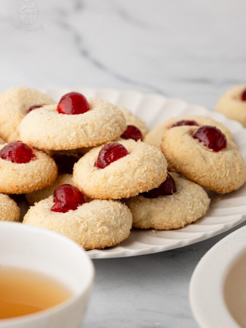 Coconut-coated Melting Moments topped with glacé cherries on a plate beside tea cups.