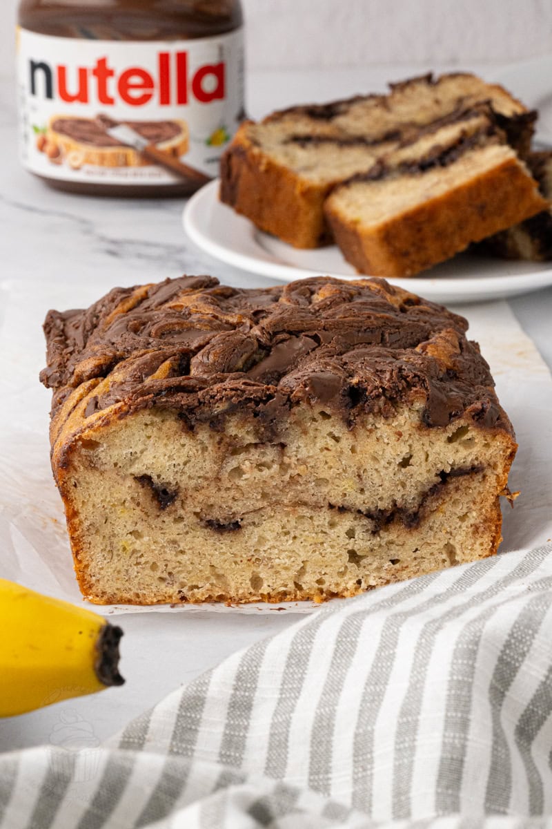 Banana Nutella loaf cake sliced on a plate with a jar of Nutella and bananas in the background.