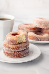 A stack of sugar-coated ring donuts on a small plate, with the top donut showing a bite taken out to reveal the fluffy texture inside.