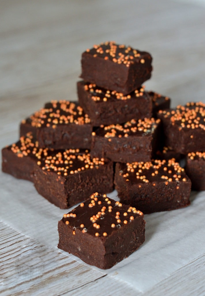 A stack of slow-cooker chocolate orange fudge, topped with small orange sprinkles and displayed on parchment paper. The background is a light, textured surface.