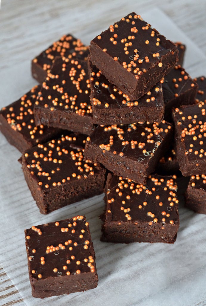 A stack of rich, sticky chocolate orange fudge topped with orange sprinkles, arranged neatly on parchment paper over a wooden surface. The vibrant sprinkles add a festive touch to the dark, fudgy goodness.