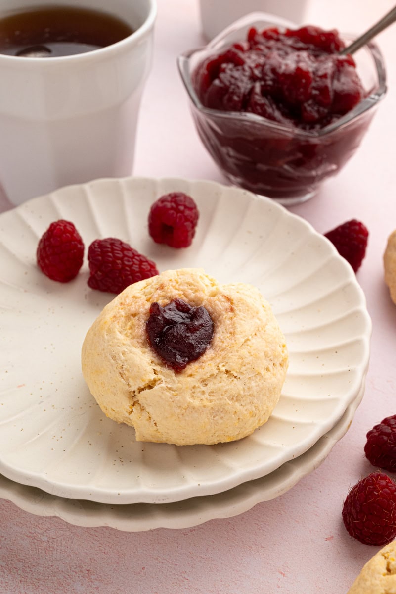 Raspberry jam-filled rasp bun served on a white plate with fresh raspberries and a cup of tea.