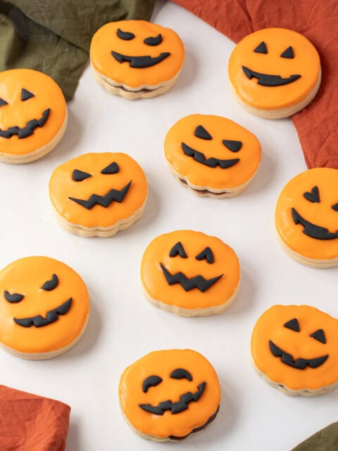 A group of orange-iced Pumpkin Empire Biscuits with black fondant pumpkin faces arranged on white surface.