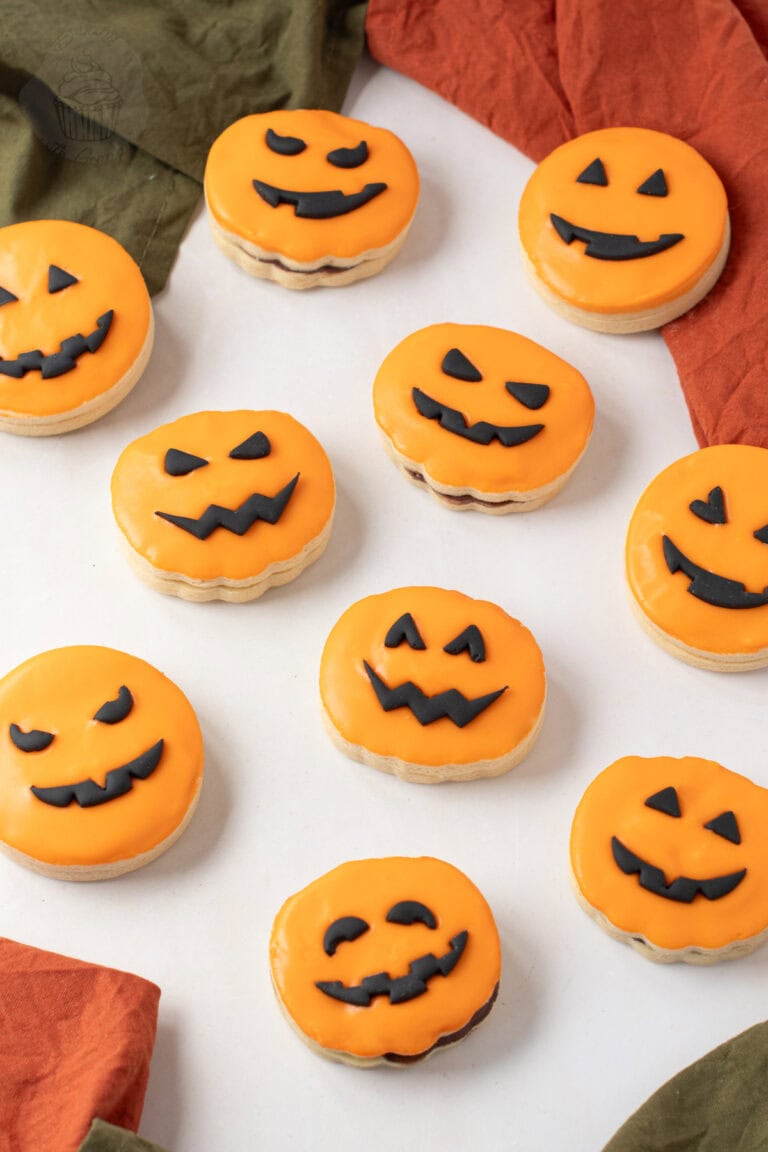 A group of orange-iced Pumpkin Empire Biscuits with black fondant pumpkin faces arranged on white surface.
