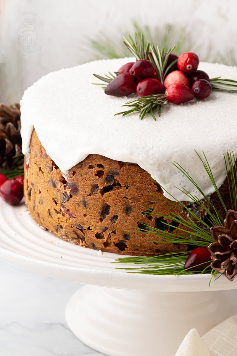 Classic British Christmas cake covered with white icing and topped with cranberries and rosemary, on a cake stand with festive greenery. Traditional UK fruit cake recipe.