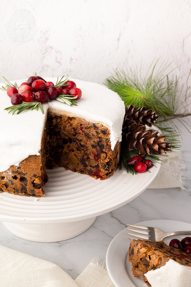 Festive British Christmas cake on a stand with one slice removed, topped with white icing, cranberries, and rosemary. Traditional UK fruit cake recipe.