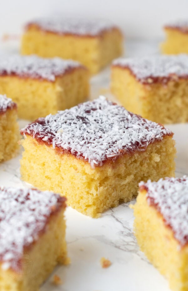 Slices of traditional jam and coconut school cake arranged on a white background, showing soft yellow sponge and coconut-covered jam topping.