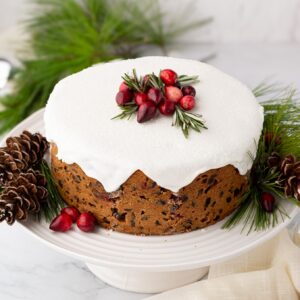 Whole Christmas cake topped with white icing, cranberries and rosemary on a cake stand.