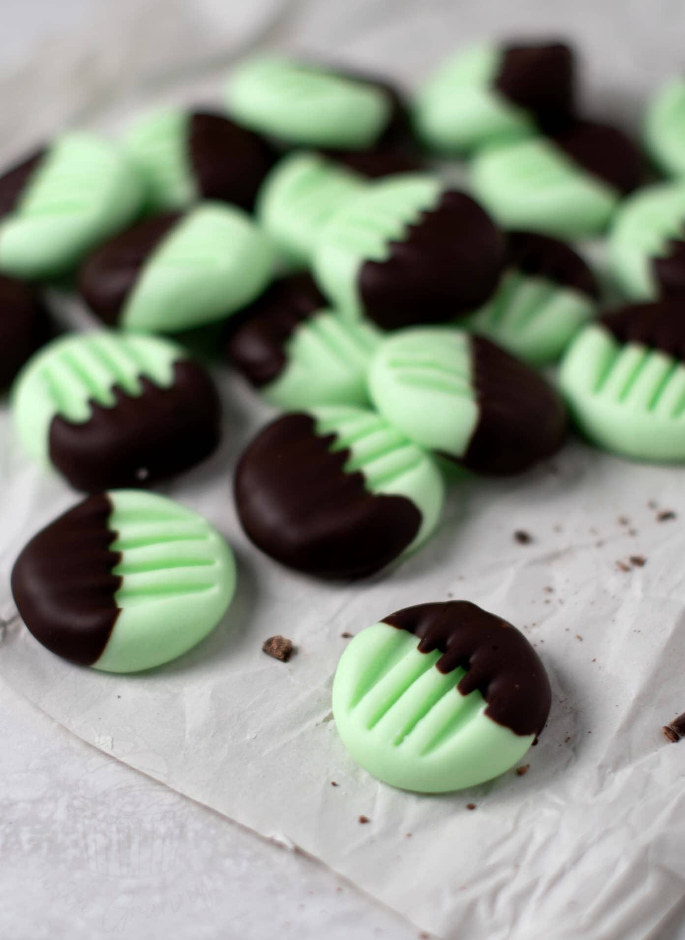 Mint chocolate peppermint creams with a green base and dark chocolate-dipped tops. Scattered on crumpled parchment paper, these peppermint fondant creams are adorned with a fork imprint pattern on the tops.