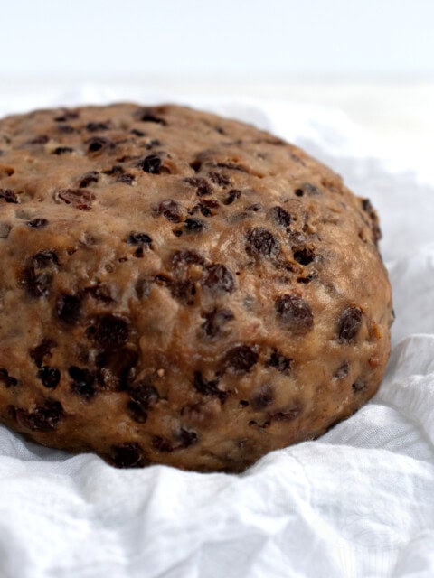 A round, dense traditional Scottish clootie dumpling, filled with visible raisins and currants, rests on a white cloth. The dumplings textured surface reflects its rich, moist composition.