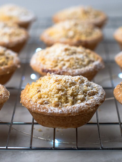 Crumble Top Mince Pies recipe from Baking with Granny. Delicious classic shortcrust mince pie, with a crumble topping, sat on top of a cooling rack, with Christmas lights twinkling underneath.