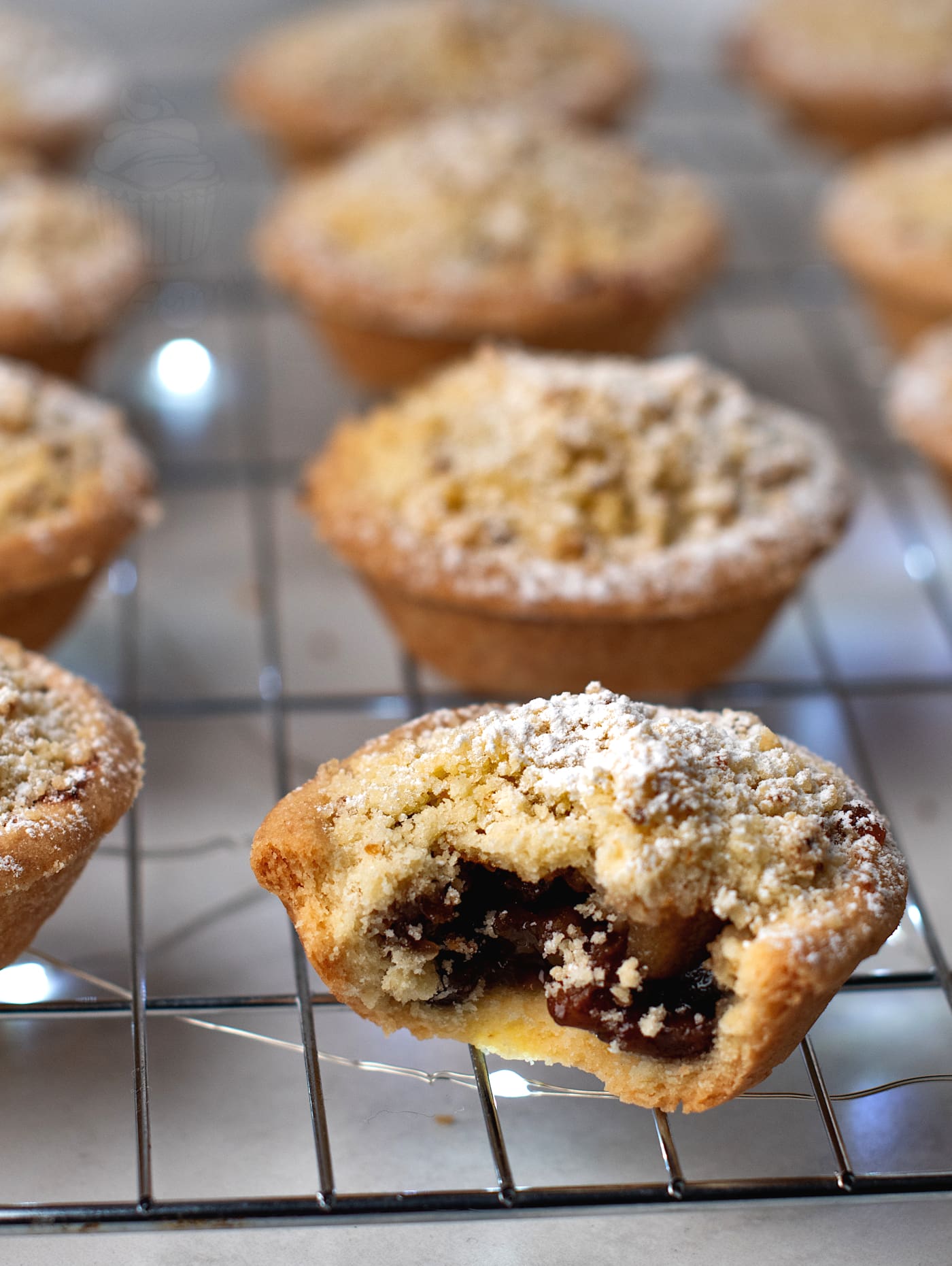 A cooling rack holds freshly baked crumble top mince pies, each dusted with powdered sugar. The focus is on one pie with a bite taken out, revealing a rich, dark fruit filling, while other whole pies sit invitingly in the background.