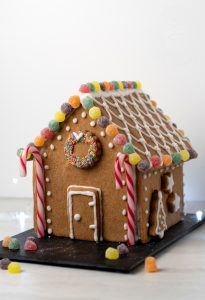 A gingerbread house crafted from a delightful UK recipe is adorned with colourful jelly tots, striped candy canes, and white icing patterns. A wreath made of sprinkles graces the front. The house rests on a black slate surrounded by scattered sweeties.