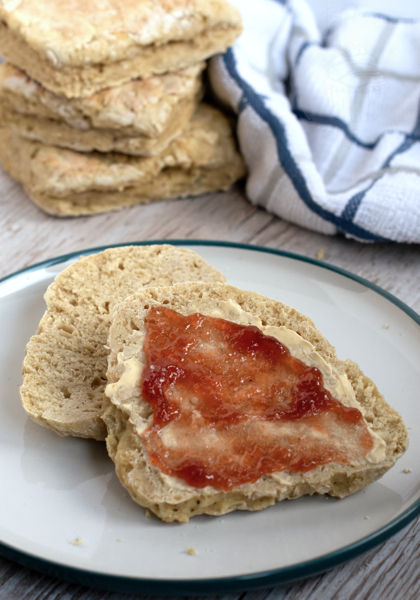 A plate with a Irish soda farl cut in half, topped with butter and strawberry jam. In the background, a stack of traditional soda farls sit next to a blue-striped white cloth on a wooden surface.