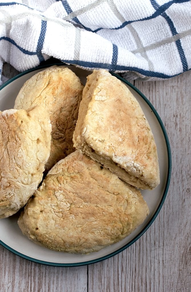 A plate of freshly baked, triangular shaped traditional Irish soda farls, sits on a wooden table. A striped white and blue kitchen towel is placed nearby.