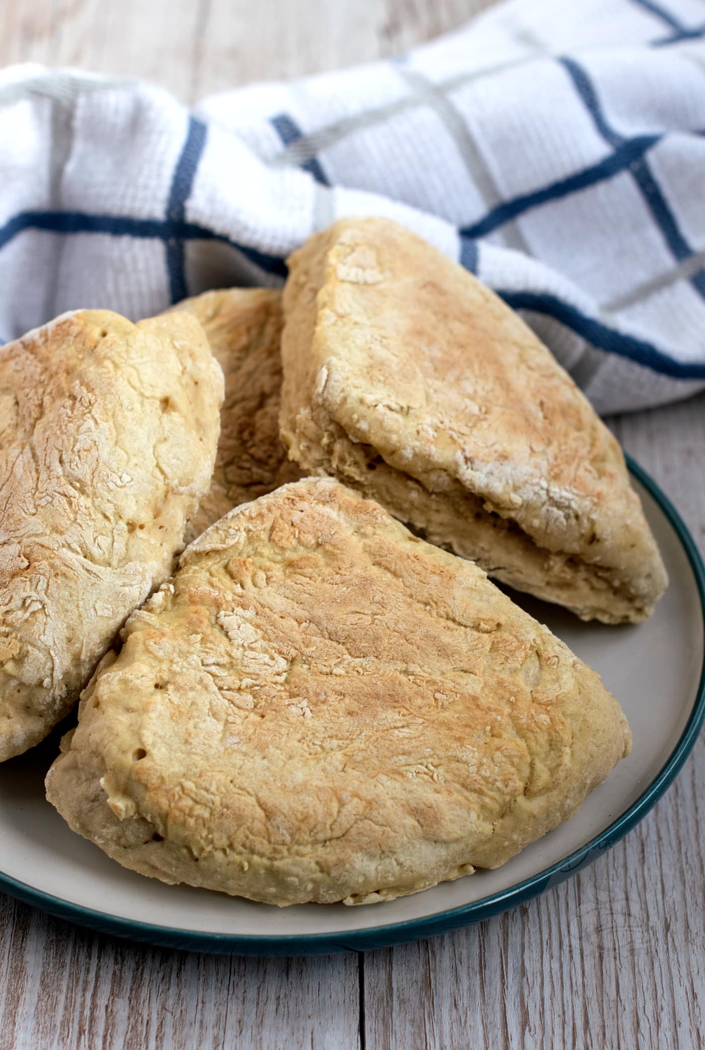 A plate of freshly baked Irish soda farls is displayed on a wooden table. The bread, made from a classic recipe, boasts a rustic, golden-brown crust. A white and blue chequered cloth provides the perfect backdrop.