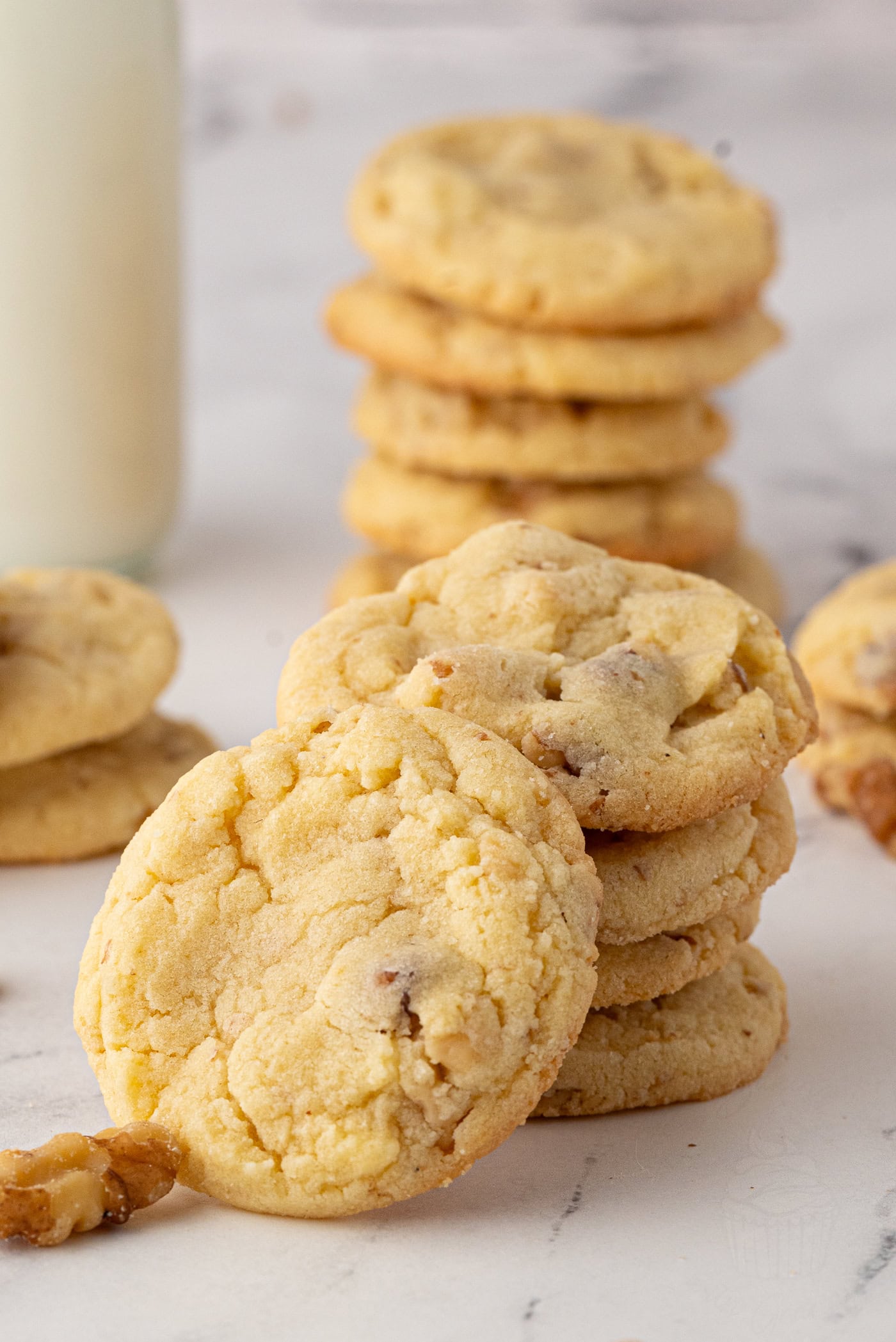 A stack of freshly baked walnut biscuits with nuts is piled high on a marble surface. In the background, more cookies are stacked with a glass of milk partially visible, creating a cosy and inviting scene, perfect for any homemade recipe.