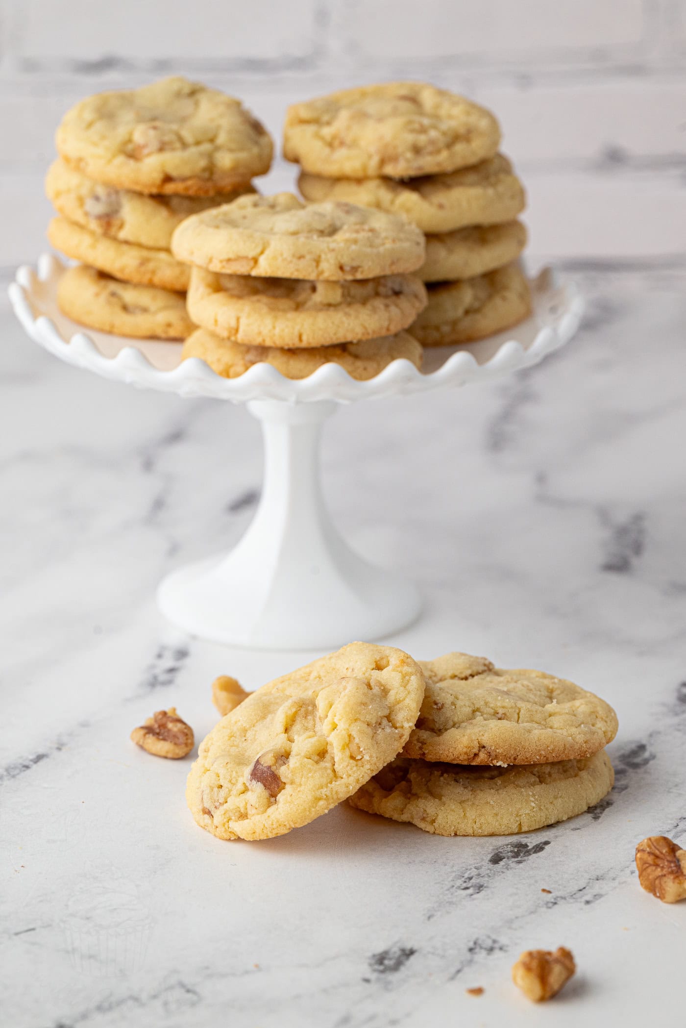 A stack of walnut cookies on a white cake stand, with three cookies and walnuts scattered on a marble surface in the foreground. The backdrop is a white brick wall.