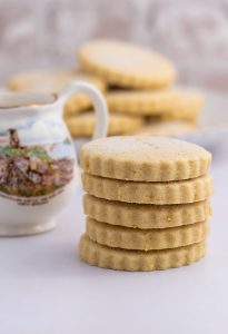 A stack of round, scalloped-edge traditional Scottish Tantallon Cake biscuits sits next to a decorative jug with a landscape design of North Berwick. More cookies are blurred in the background, all on a light surface, evoking the charm of an age-old recipe.