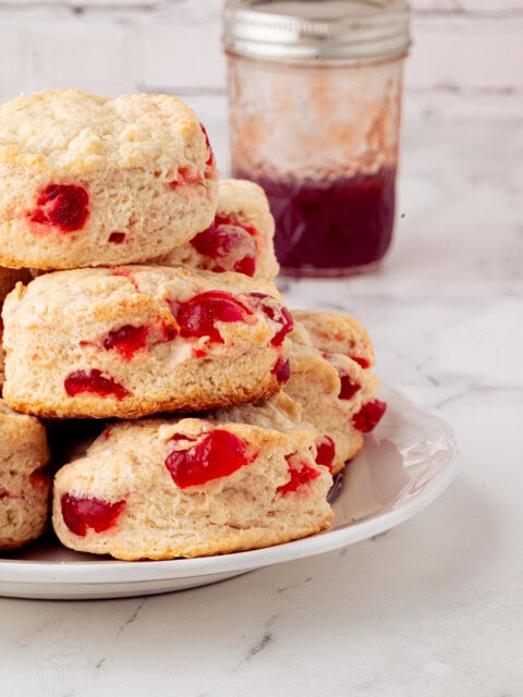 A stack of cherry scones, showcasing their delightful recipe with visible fruit pieces, sits on a white plate. In the background, a glass jar brimming with red jam complements the scene on a sleek marble countertop against a partially visible brick wall.