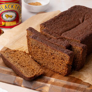 Sliced gingerbread loaf rests on a wooden cutting board, its deep brown hues inviting. Nearby, a can of black treacle and a small white dish with brown sugar add to the scene, accompanied by a knife and plate. The loaf is carefully placed on parchment paper.