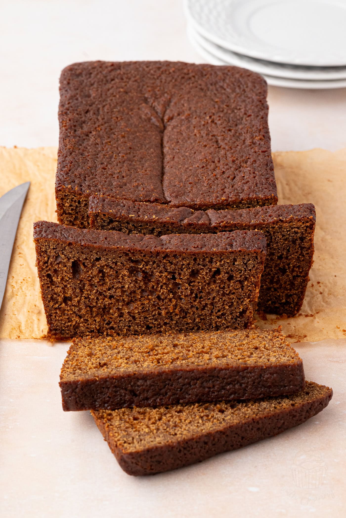 A sliced gingerbread loaf rests on parchment paper, a knife beside it. It has a dark brown hue and is dense in texture. In the background, stacked white plates await their moment.