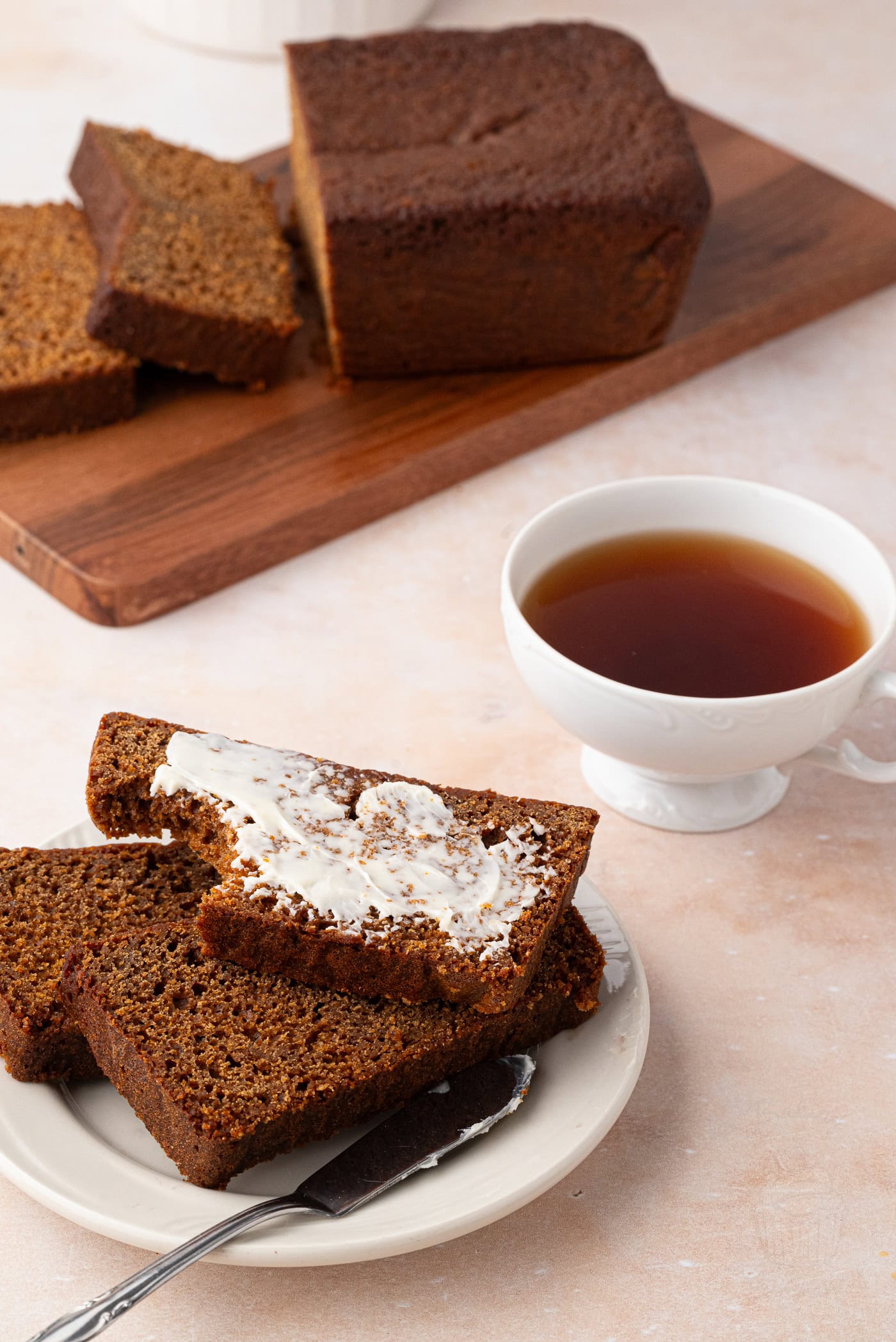 Slices of gingerbread cake sit on a white plate, with one slice topped with butter. A butter knife rests beside them. In the background, more gingerbread and a hint of the gingerbread cake sitting on a wooden board. A cup of tea is placed nearby on a light-coloured surface.