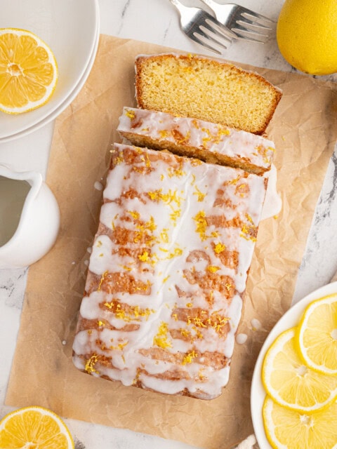 A lemon drizzle loaf cake with icing and lemon zest on top sits on parchment paper, embodying the perfect recipe. Slices of lemon surround the cake. Nearby are a pitcher, two forks, plates, and a whole lemon on a marble surface.