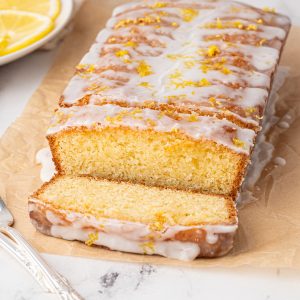 A lemon drizzle cake recipe unfolds with a lemon loaf adorned in white icing and zest, partially sliced. Resting on parchment, surrounded by fresh lemon slices and accompanied by a fork, the scene is elegantly set on a marble surface.