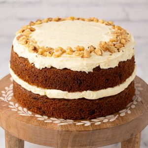 Two-layer carrot cake topped with buttercream and chopped walnuts, displayed on a wooden cake stand with inlaid detail.