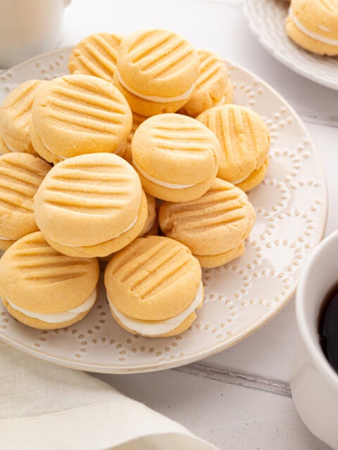 Overhead view of custard cream biscuits arranged on a cream plate beside white coffee mugs and napkins.