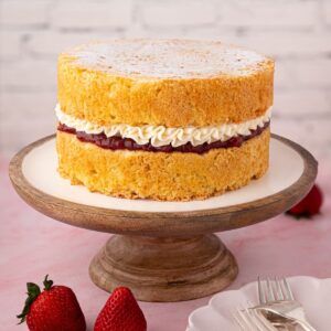 Victoria Sponge filled with strawberry jam and buttercream on a wooden cake stand.