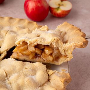 Slice of apple pie on a white plate beside fresh apples and the whole pie.