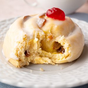 Side view of a Belgian bun with a bite taken out, topped with white icing and a red glacé cherry, served with tea.