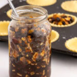 Homemade Christmas mincemeat in a glass jar beside unbaked mince pies in pastry cases.