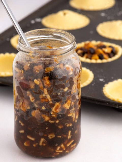 Homemade Christmas mincemeat in a glass jar beside unbaked mince pies in pastry cases.