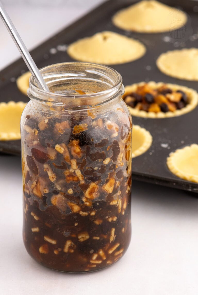 Homemade Christmas mincemeat in a glass jar beside unbaked mince pies in pastry cases.