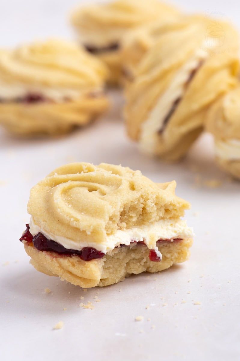 Part-eaten homemade Viennese Whirl cookies filled with raspberry jam and vanilla buttercream, with more biscuits in the background.