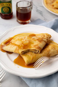 Two folded pancakes drenched in rich golden syrup on a white plate, with a silver fork resting on the side.