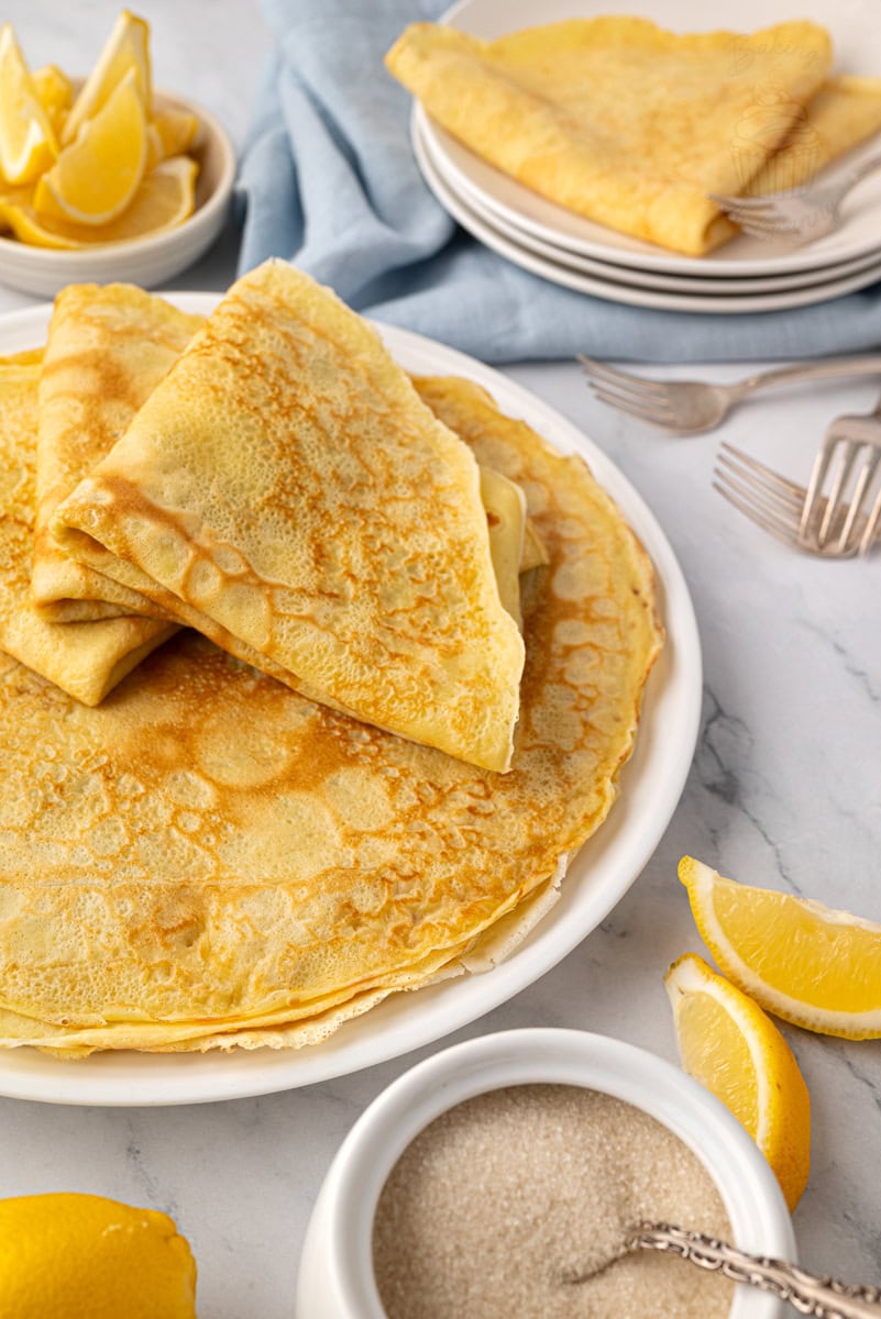 Plate of homemade British pancakes with caster sugar and lemon slices on a marble surface.