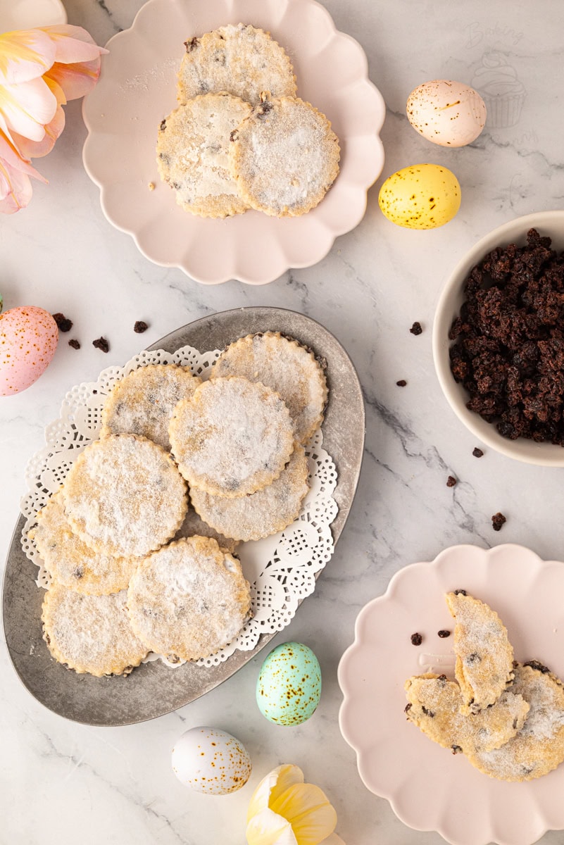 Overhead view of Easter biscuits on serving plates with currants, surrounded by pastel Easter eggs and baking ingredients.