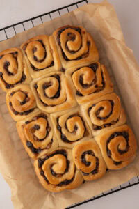 Freshly baked Chelsea Buns cooling on baking paper over a wire rack.