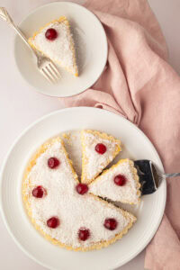 Whole Manchester Tart cut into slices and topped with desiccated coconut and glace cherries on a serving plate.