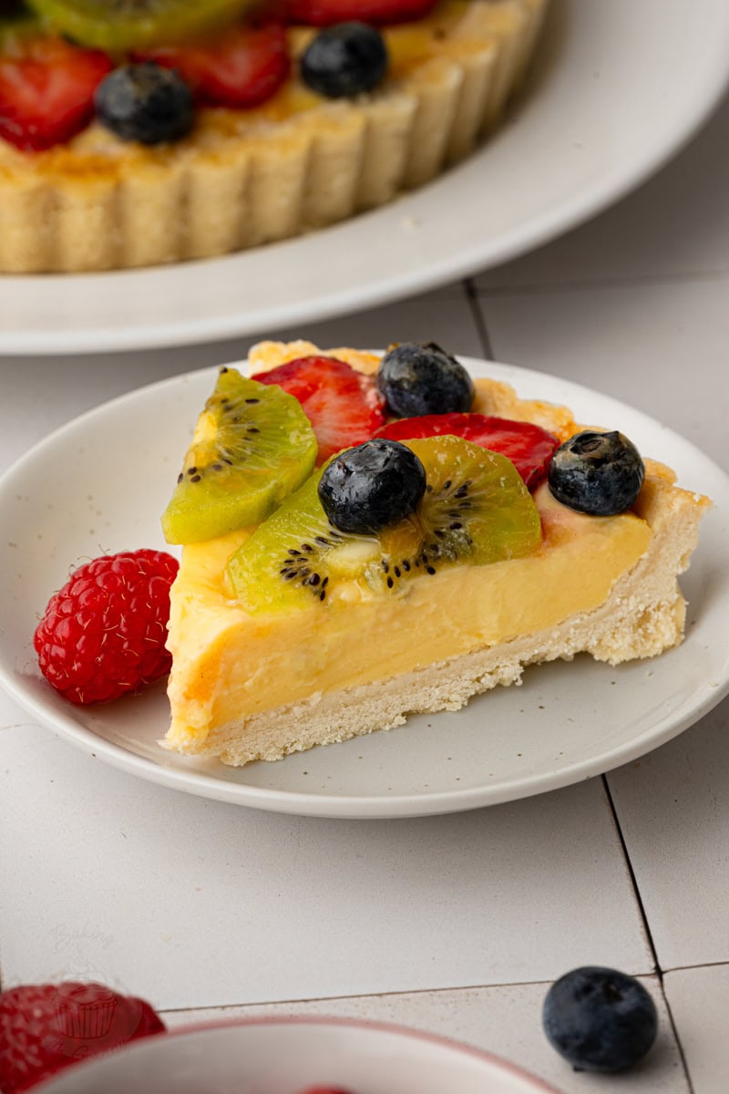 A side profile view of a fruit tart slice showing the contrast between the thick, creamy custard layer and the flaky baked tart crust, garnished with fresh berries and a glossy apricot glaze.