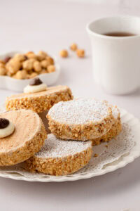 Eye-level close-up of a stacked pile of icing-sugar-dusted Japonaise cakes on a white plate with a vintage lace doily.