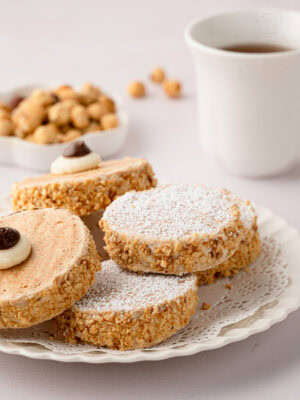 Eye-level close-up of a stacked pile of icing-sugar-dusted Japonaise cakes on a white plate with a vintage lace doily.